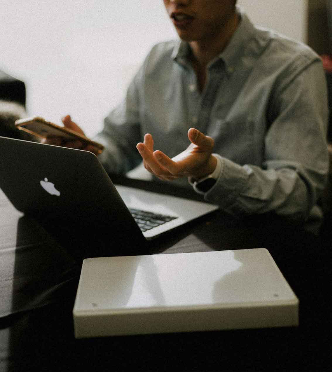 Person in a button-up shirt gesturing with one hand while holding a smartphone near an open MacBook on a dark table.