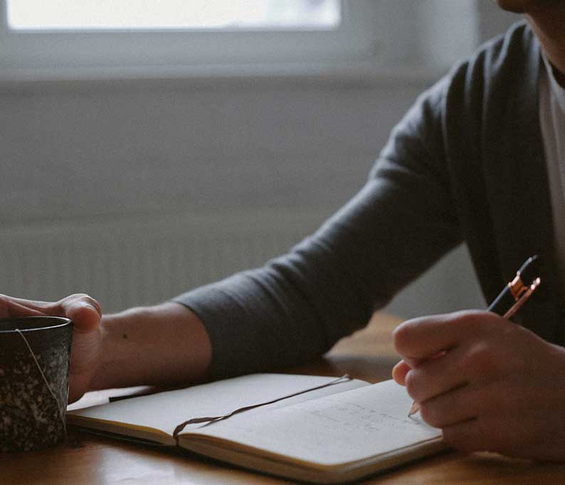 Person writing in an open notebook at a wooden table while holding a mug with a tea bag string visible.