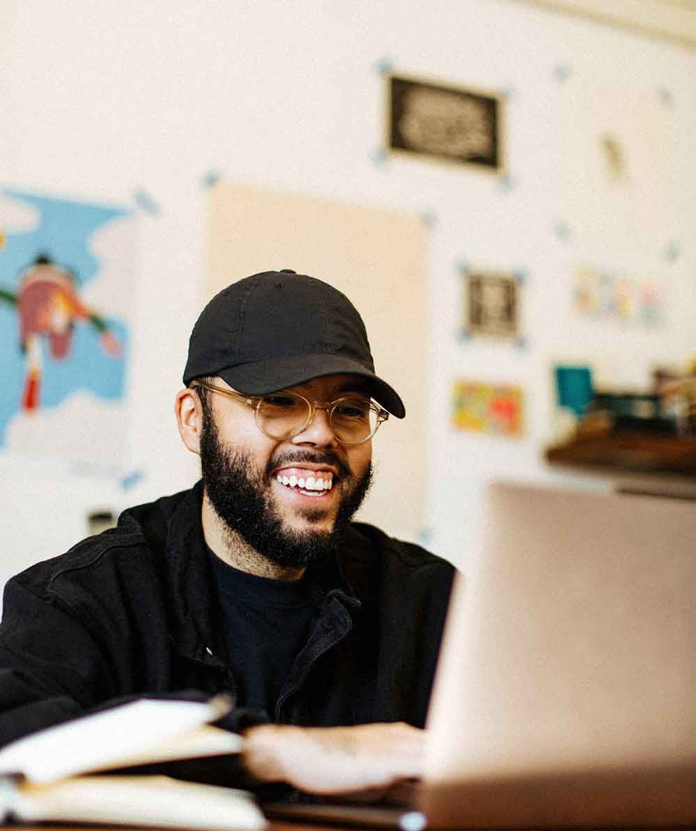 Smiling man with glasses and a black cap working on a laptop at a desk with notebooks.