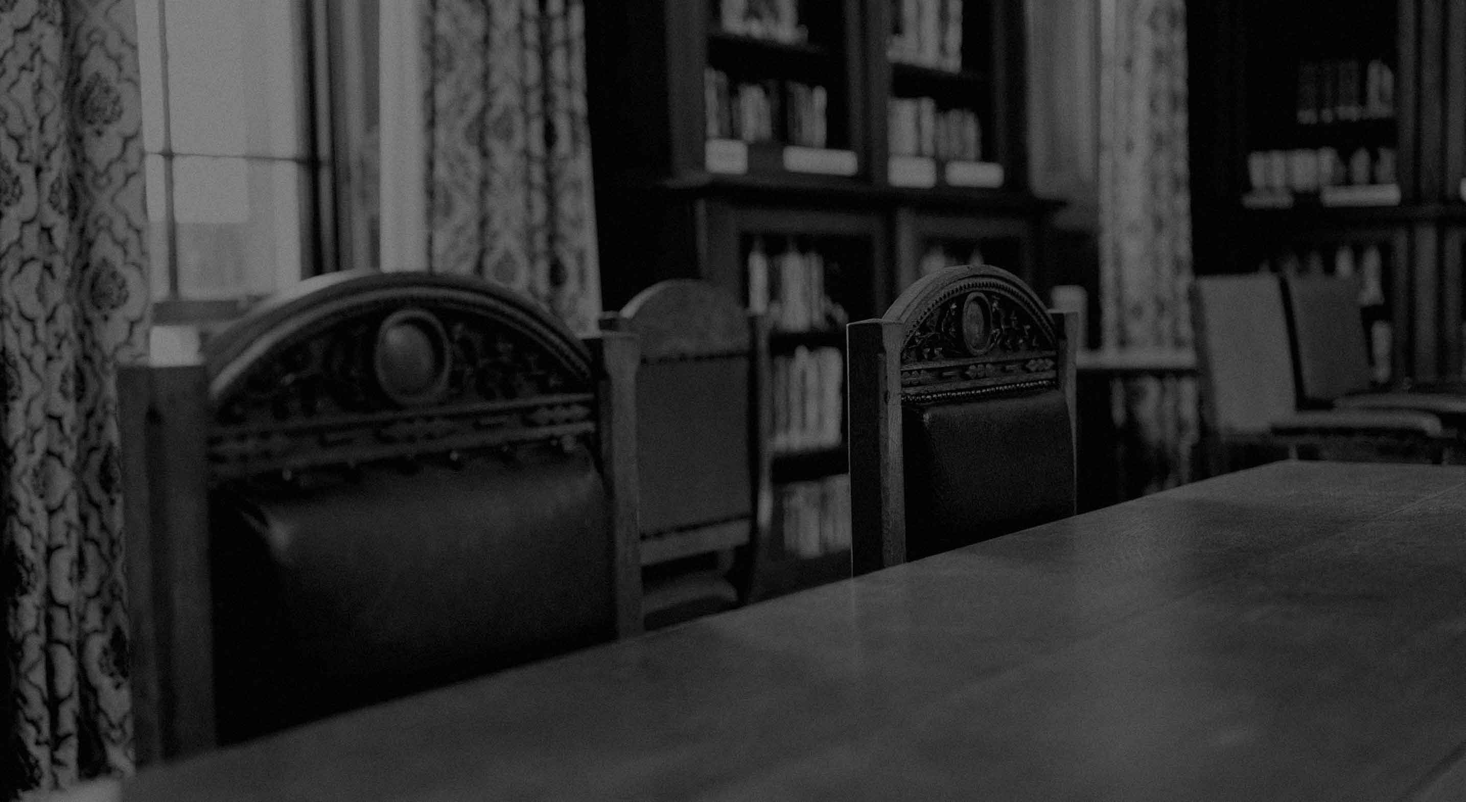 Empty ornate wooden chairs with leather backs around a wooden table in a dimly lit library room with bookshelves and patterned curtains.