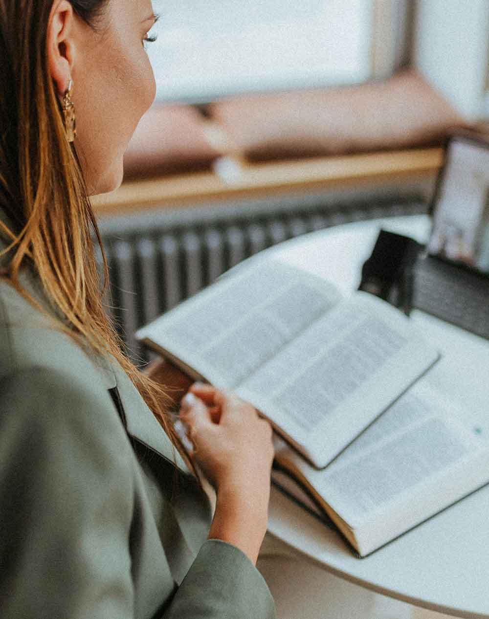 Woman with long brown hair wearing a green blazer reading an open book at a round table near a window.