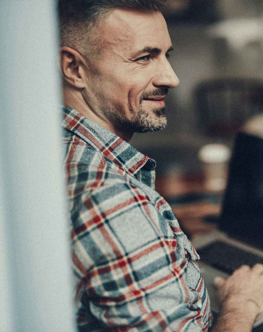Smiling man with short hair and beard wearing a plaid shirt, sitting indoors near a window.