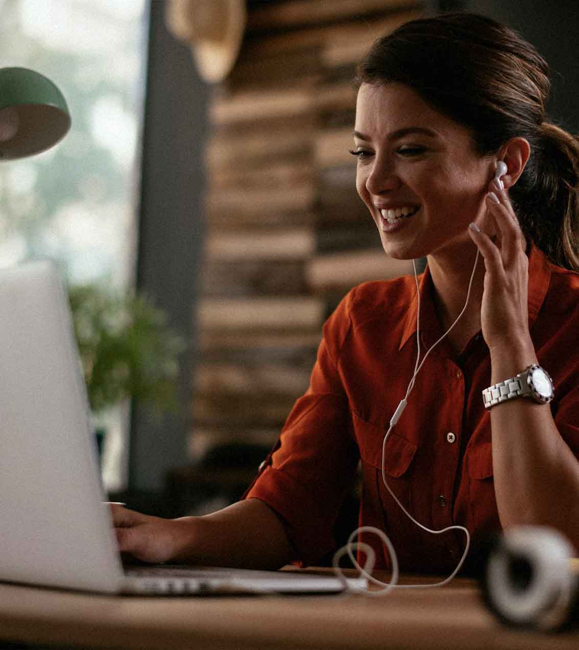 Smiling woman with dark hair wearing earphones and a watch, sitting at a table using a laptop.