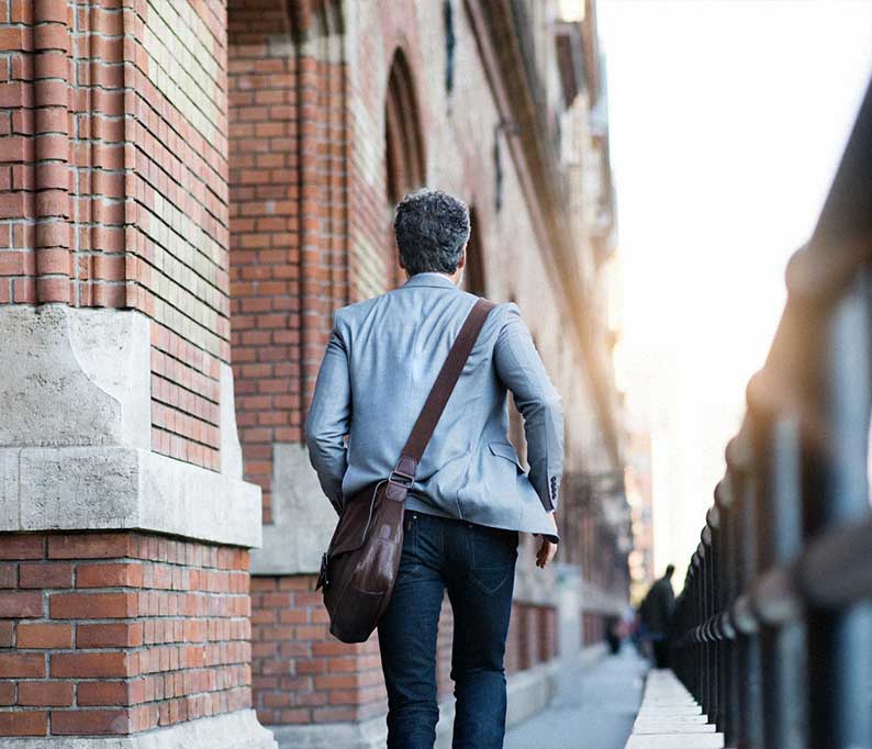 Man in a gray blazer walks along a sidewalk next to a brick building with a brown shoulder bag.