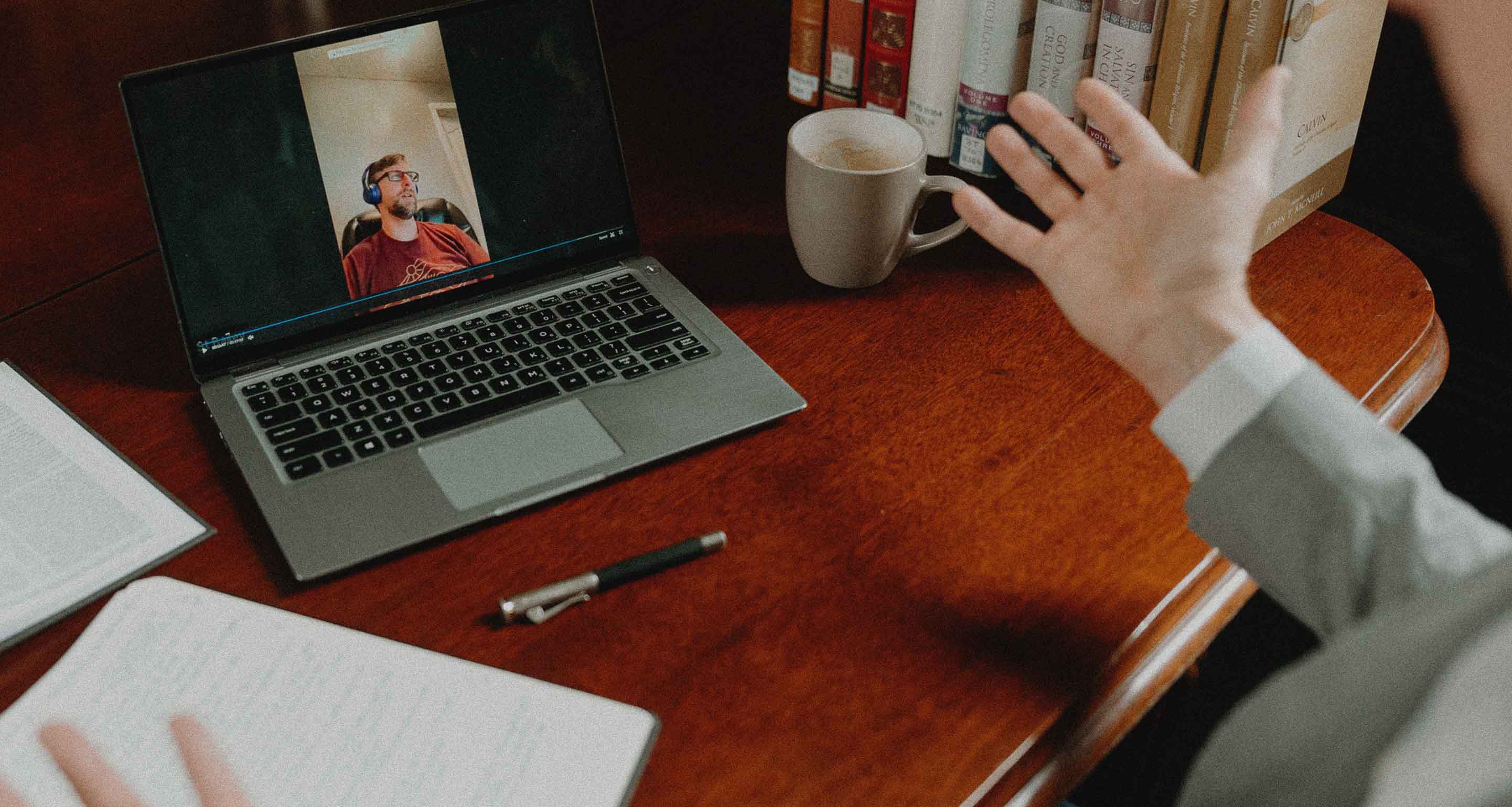 Person gesturing toward a laptop screen showing a video call with a man wearing headphones, on a wooden table with books, a coffee cup, a pen, and open notebooks.