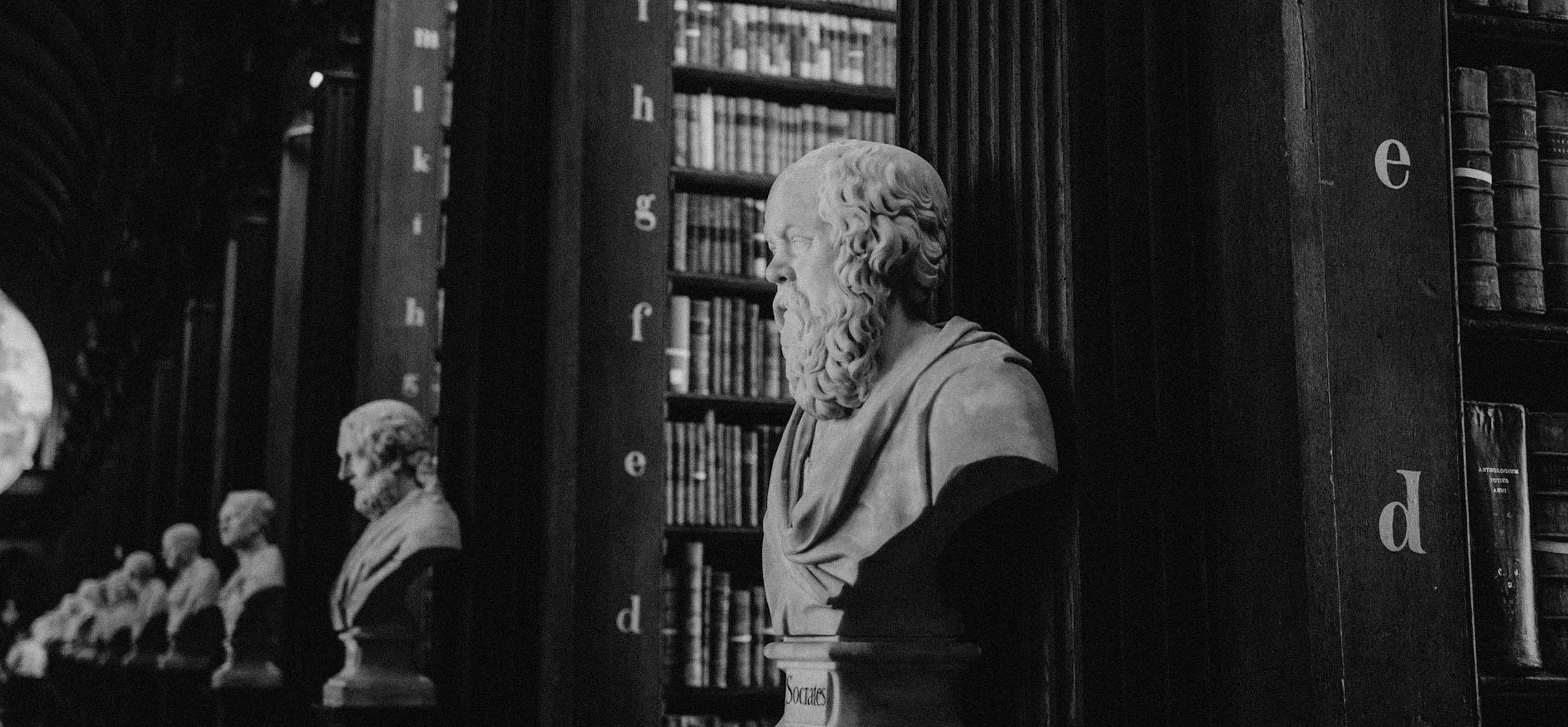 Black and white photo of a row of classical bust statues, with the closest labeled Socrates, against a background of bookshelves.