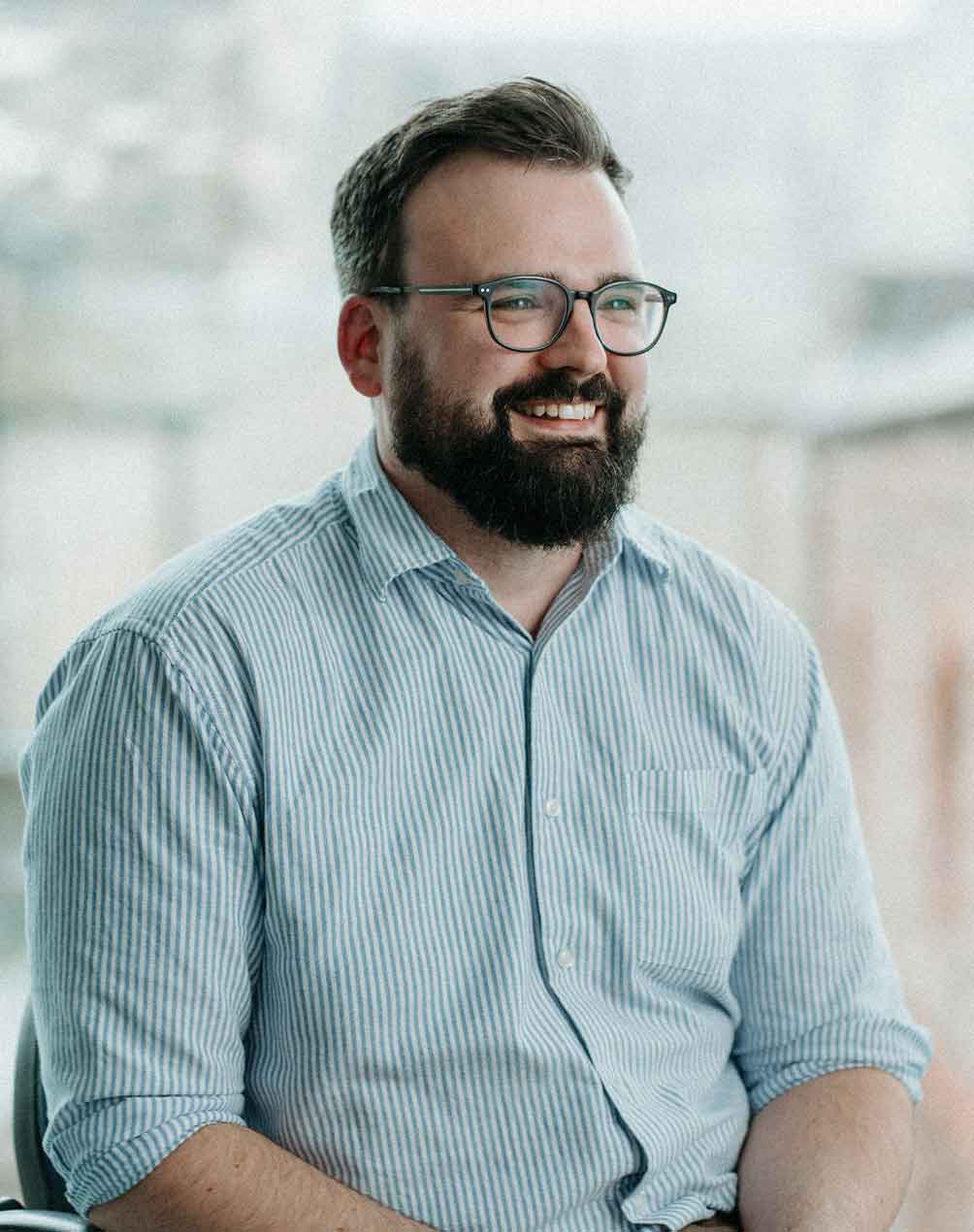 Smiling man with glasses and beard wearing a light blue and white striped shirt.