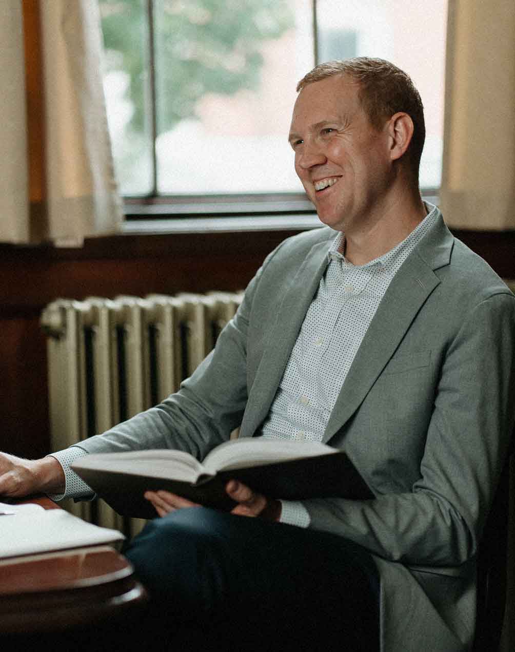 Smiling man in a light gray blazer holding an open book while sitting near a window.