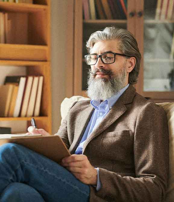 Bearded man wearing glasses, a brown blazer, and blue shirt, sitting on a couch writing on a clipboard.