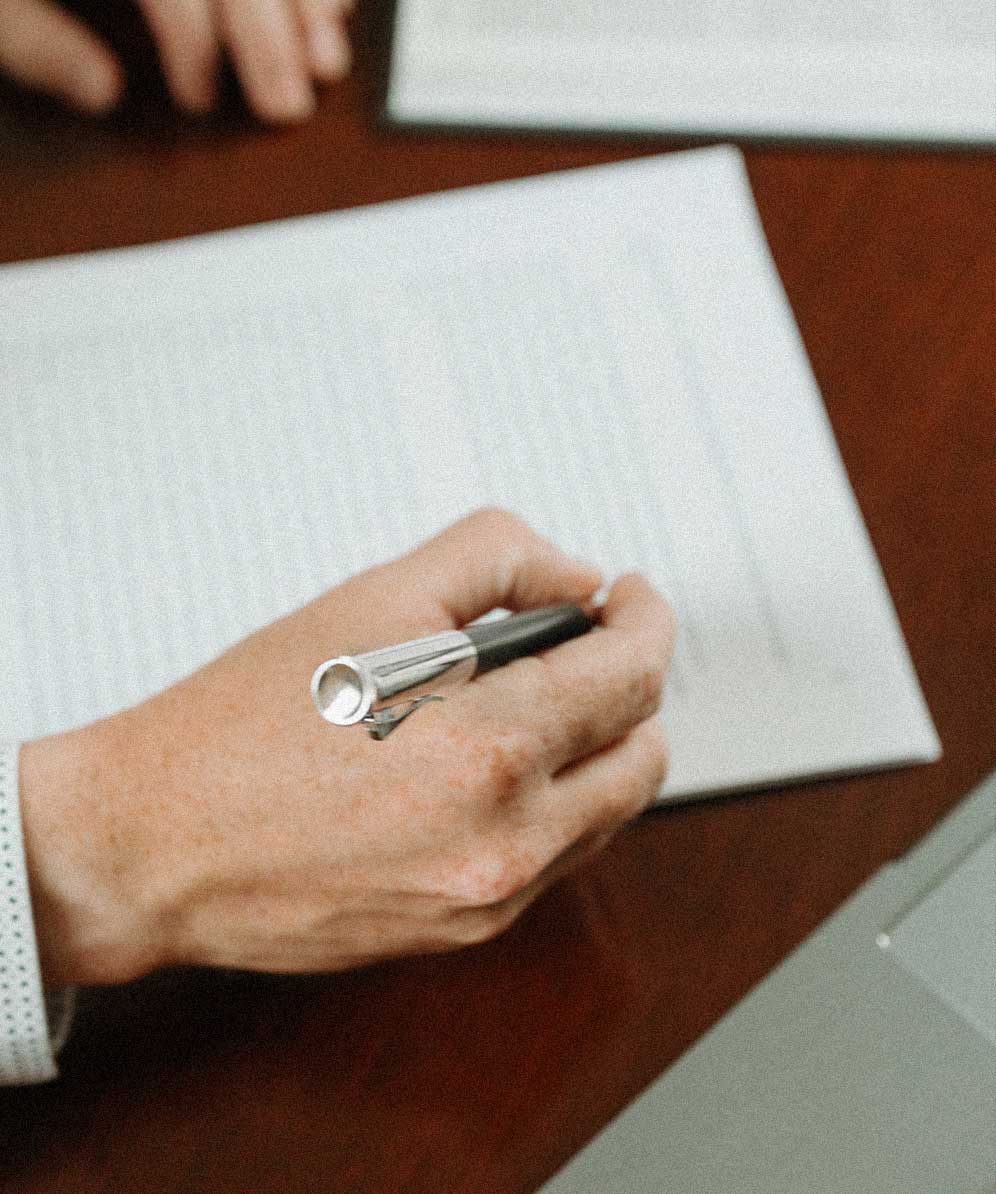 Close-up of a hand holding a pen above an open notebook on a wooden desk with a laptop partially visible.