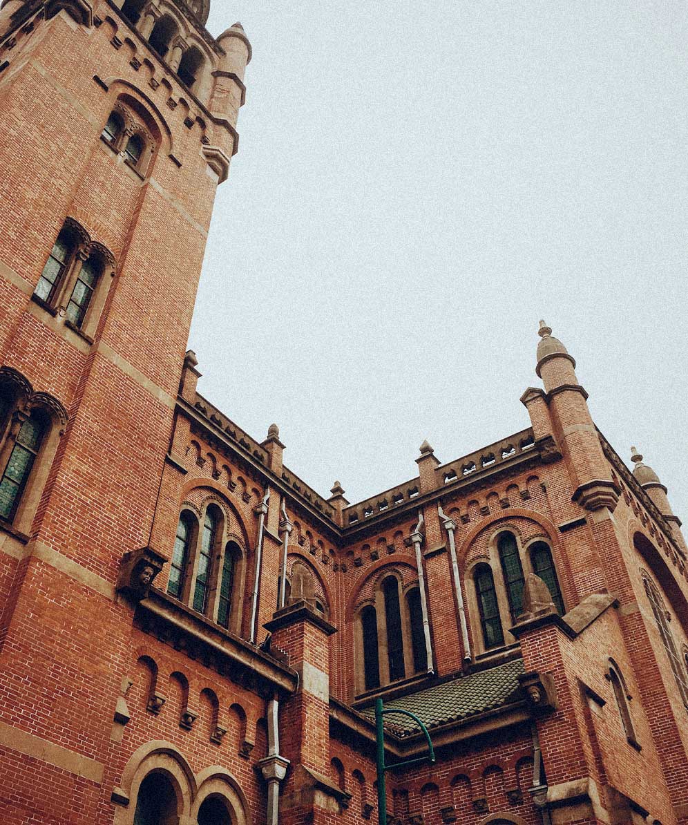 Low-angle view of a red brick Gothic-style building with arched windows and decorative stone elements under an overcast sky.