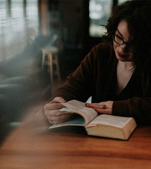 Woman with glasses reading and turning pages of a book at a wooden table in a dimly lit room.