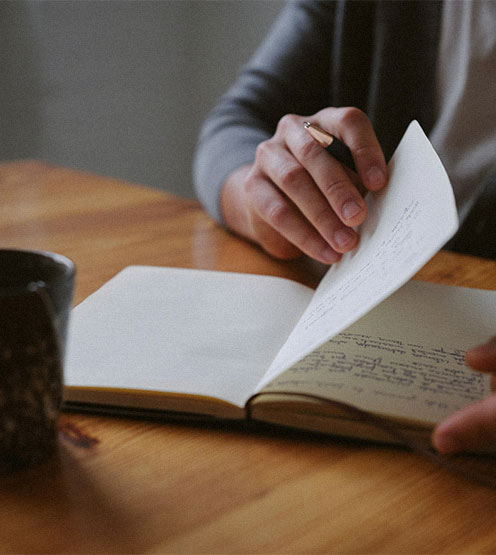 Person turning a page in a notebook while holding a pen on a wooden table next to a dark patterned cup.