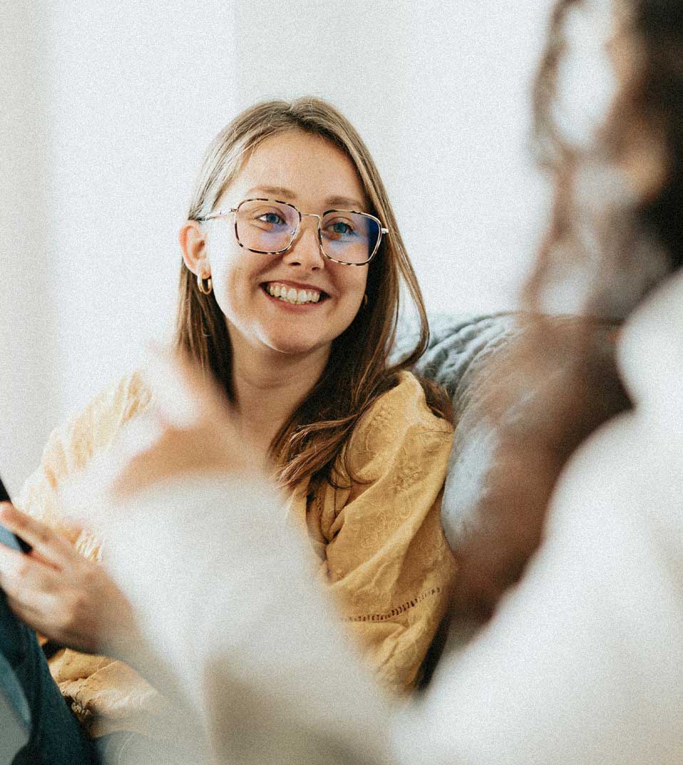 Smiling woman with glasses and a yellow sweater, holding a phone and interacting with another person.