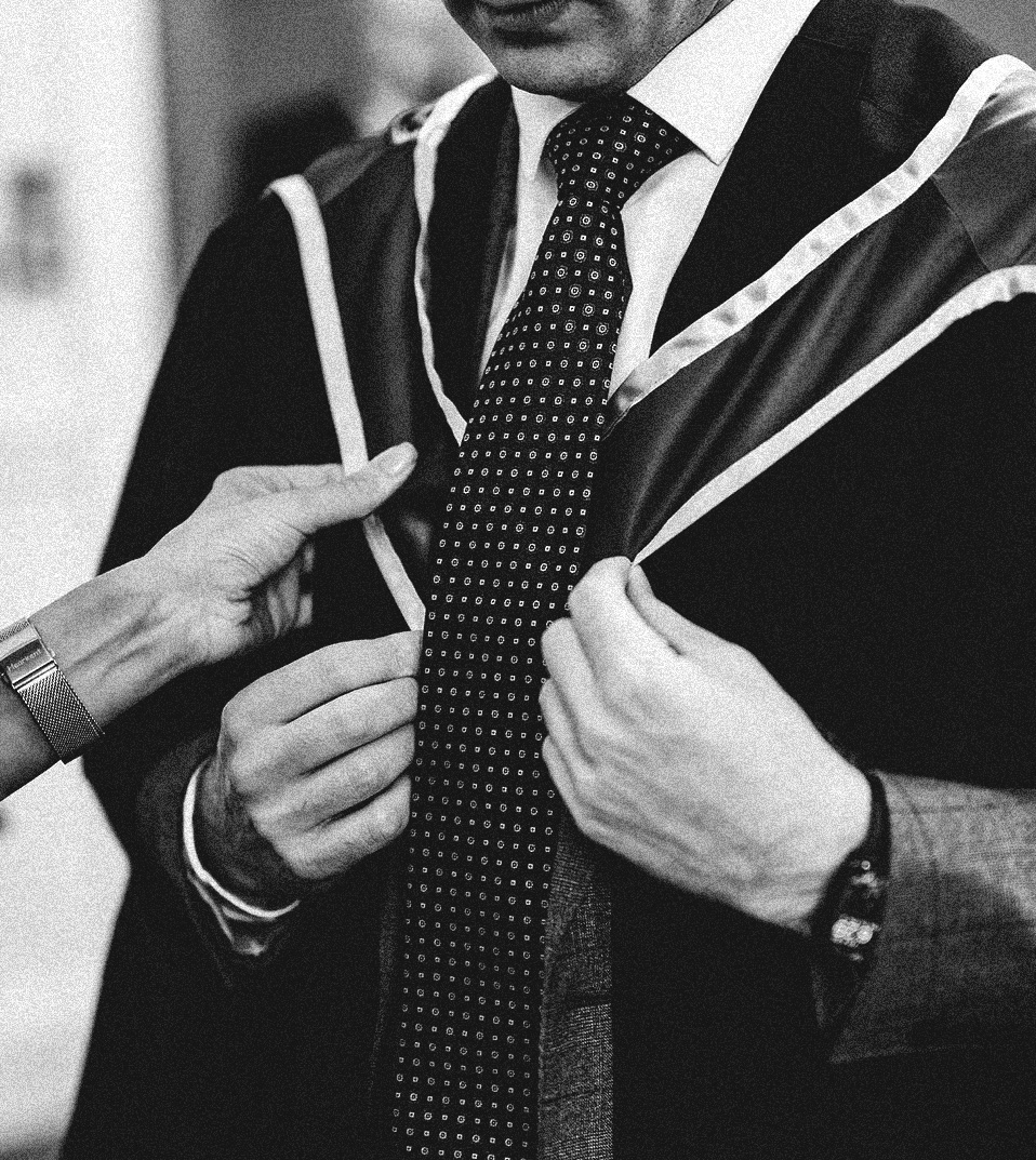 Close-up of a graduate from The Alexandrian Institute adjusting his academic gown over his suit and patterned tie. This photo was taken at a graduation ceremony at Union Theological College, Belfast.