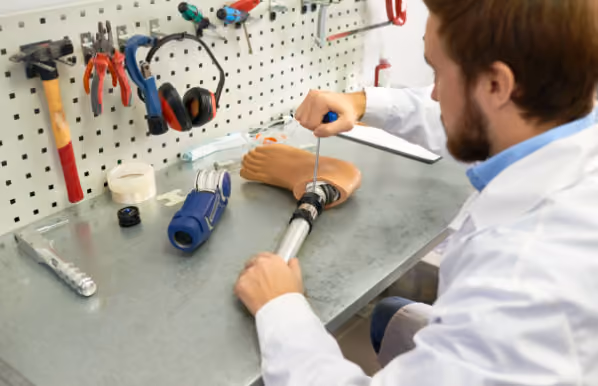 A man in a white lab coat working on assembling a prosthetic leg at a workbench with tools hanging on a pegboard and other prosthetic parts on the table.