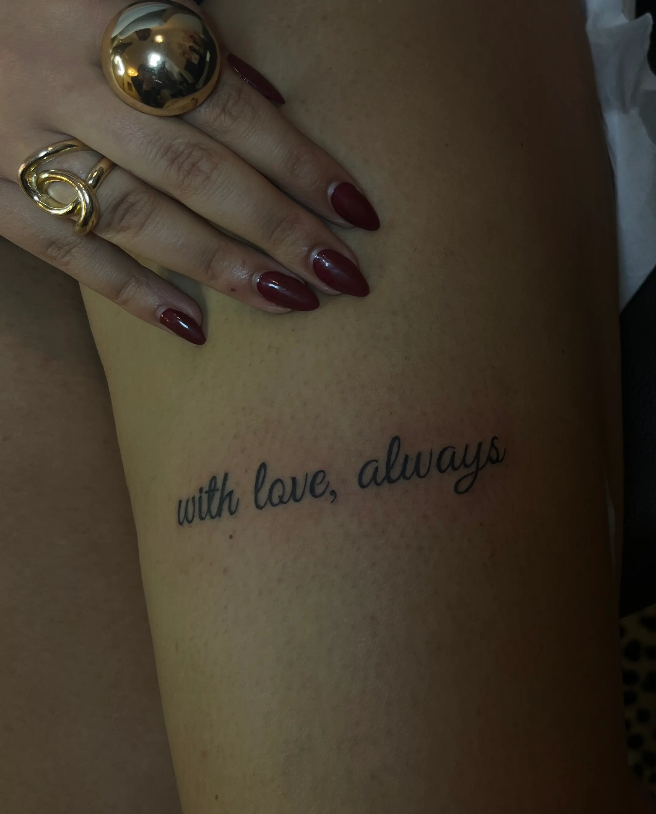 Frisch gestochenes Schriftzug-Tattoo mit rotem Nageldesign und goldenen Ringen am Finger.