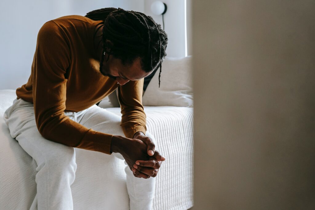 A man on the edge of a bed, looking down as if struggling to sleep, in pain or stressed