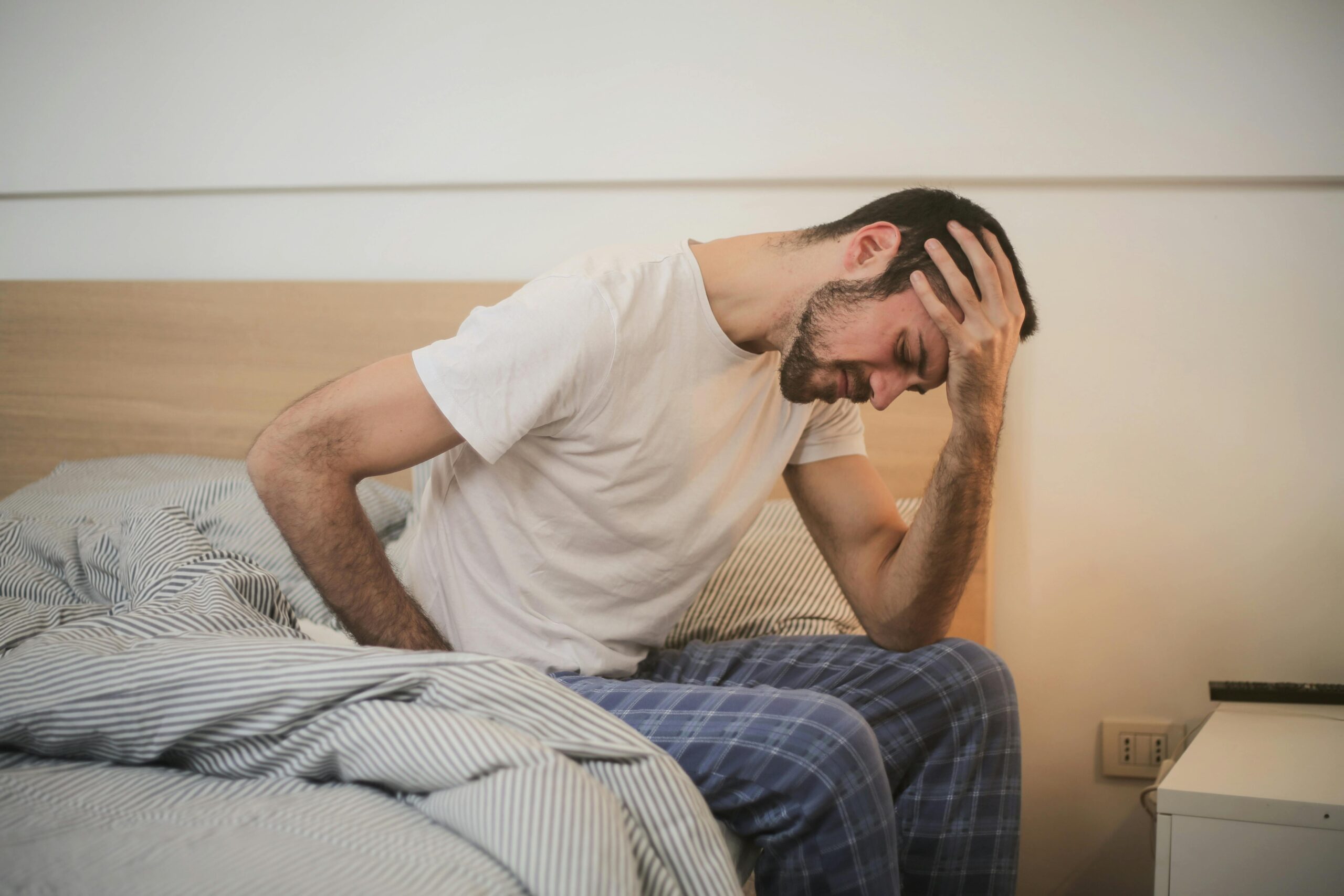 A man sits on the side of his bed with his hand on his head, suffering from a migraine.