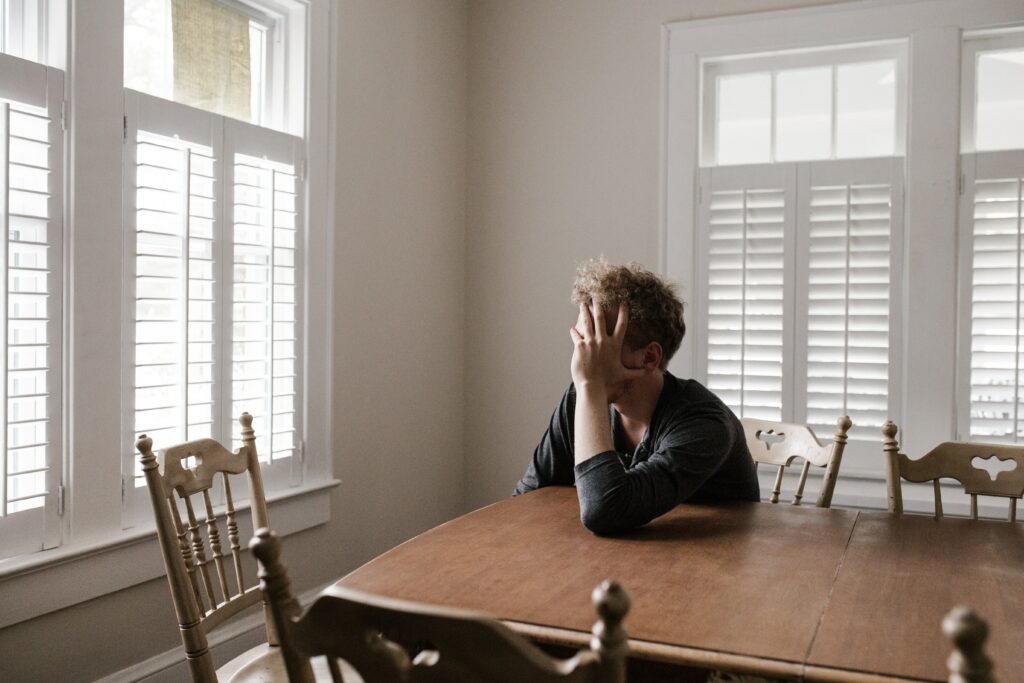 A man sat at a dining table with his head in one hand, looking out the window as if in pain