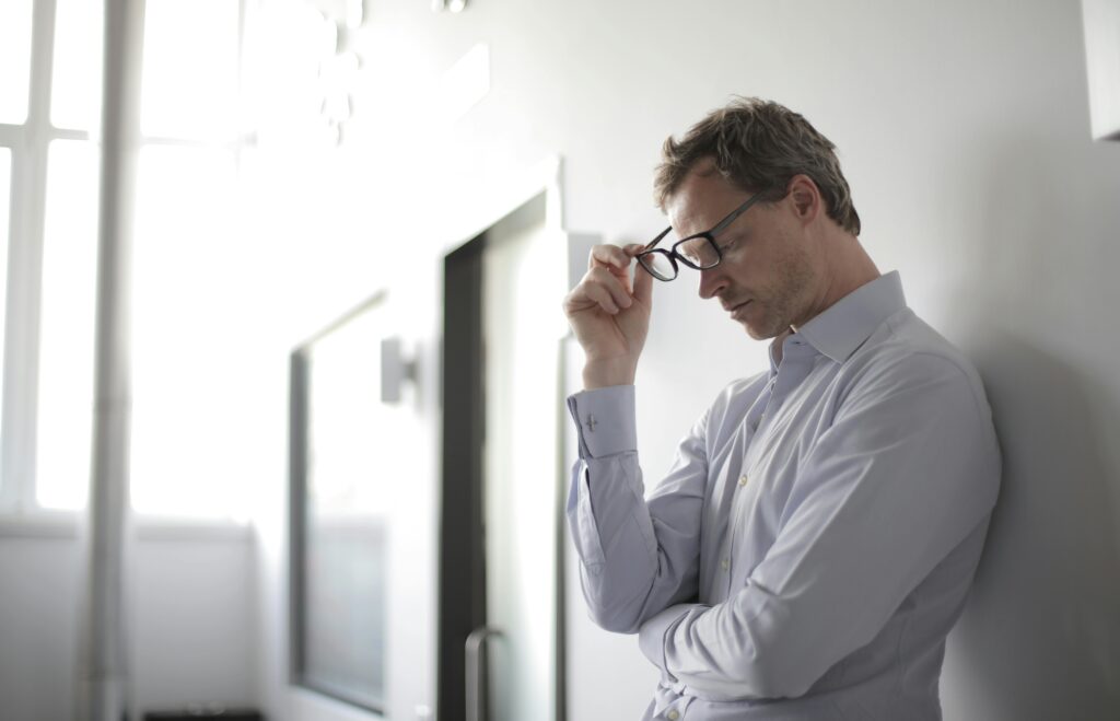 A man leans with his back against the wall, as he takes off his glasses and looks at the ground