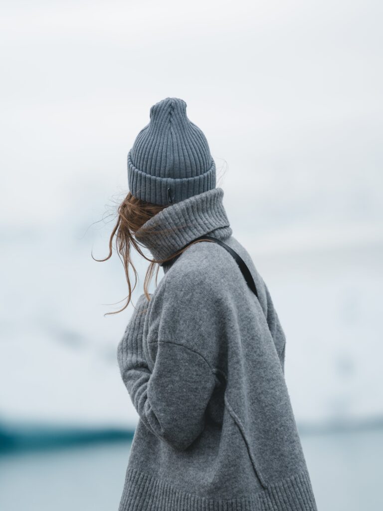 A woman wearing an oversized cardigan and beanie, looking out over the sea