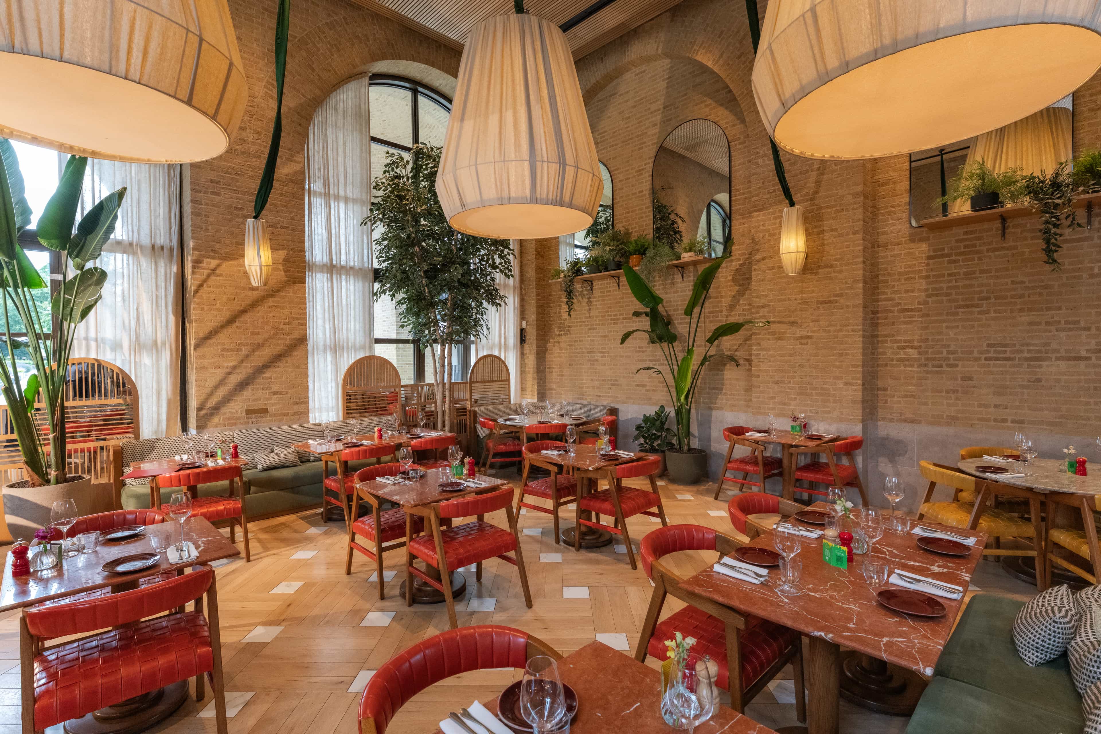 Bright and stylish restaurant interior with high arched windows, warm brick walls, large fabric pendant lights, wooden tables, red leather chairs, and lush indoor plants creating a cozy and elegant atmosphere