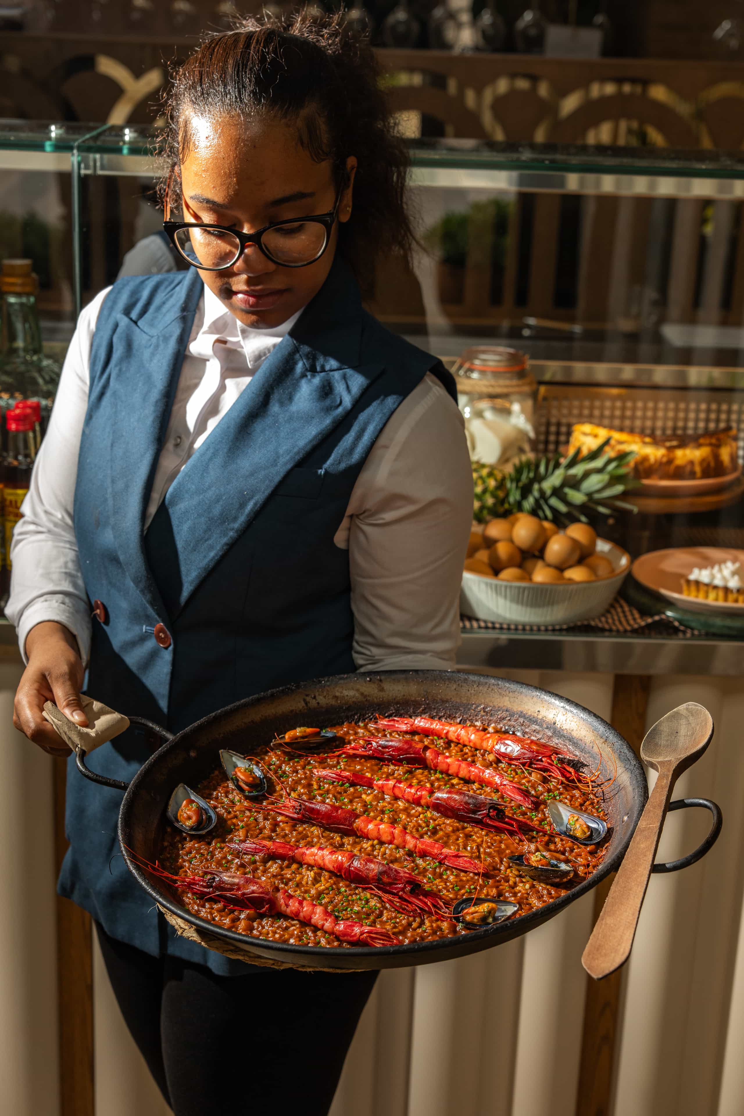 Waitress in a navy blue vest holding a large paella pan with prawns and mussels, standing in front of a counter with eggs, fruits, and jars in a warmly lit restaurant setting