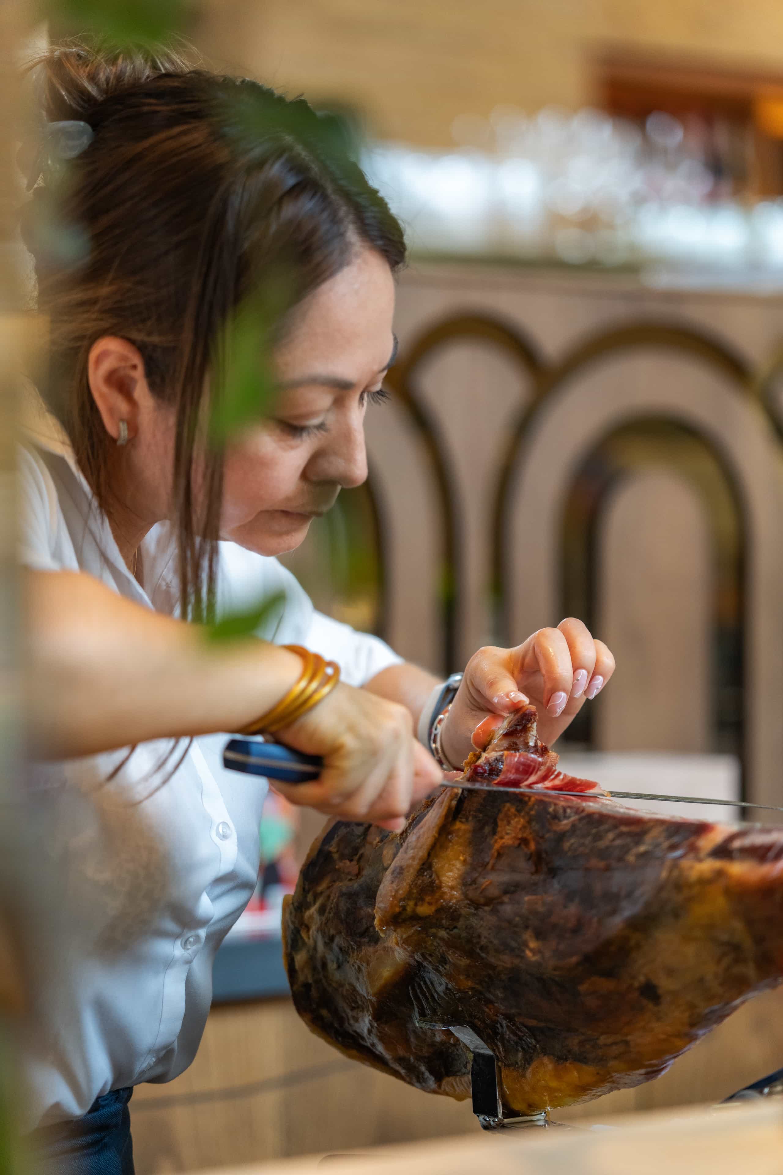 Close-up of a woman skillfully slicing cured ham from a jamón ibérico leg with concentration, in an elegant restaurant setting.