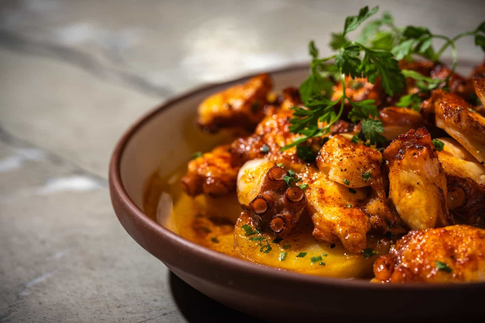 Close-up of a plate of cured meat slices topped with thin shavings of cheese, dollops of herb sauce, and microgreens, served on a white plate with a brown-rimmed edge