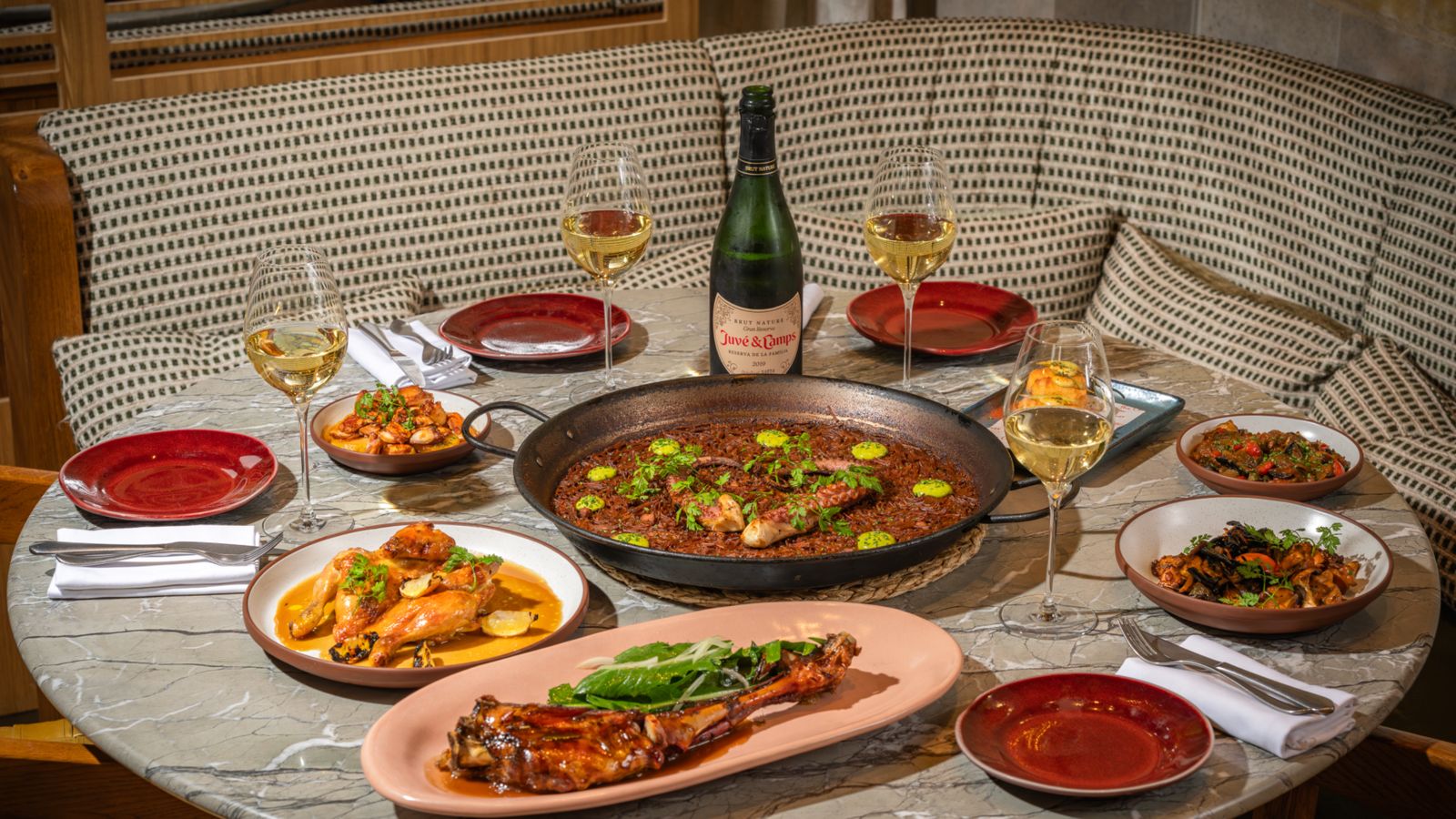 Outdoor table setting with a bottle of white wine and two glasses, framed by greenery and a historic brick building in the background