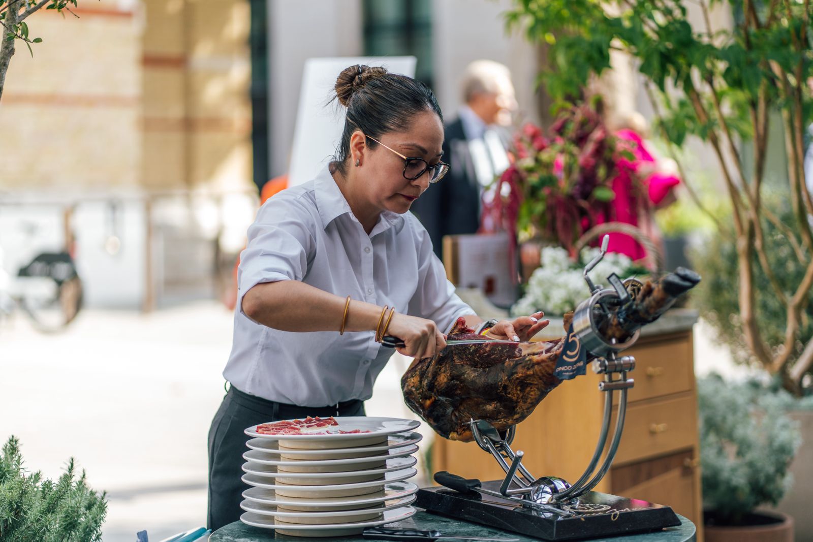 Sunlit indoor dining space with patterned benches, wooden tables, and an olive tree surrounded by lush greenery