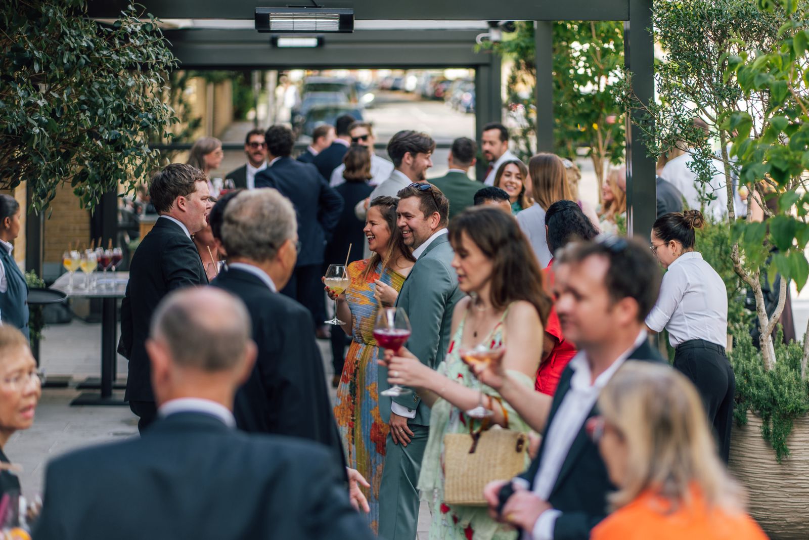 Exterior view of The Campaner with large arched windows, red terrace seating, and potted trees lining a wide stone plaza