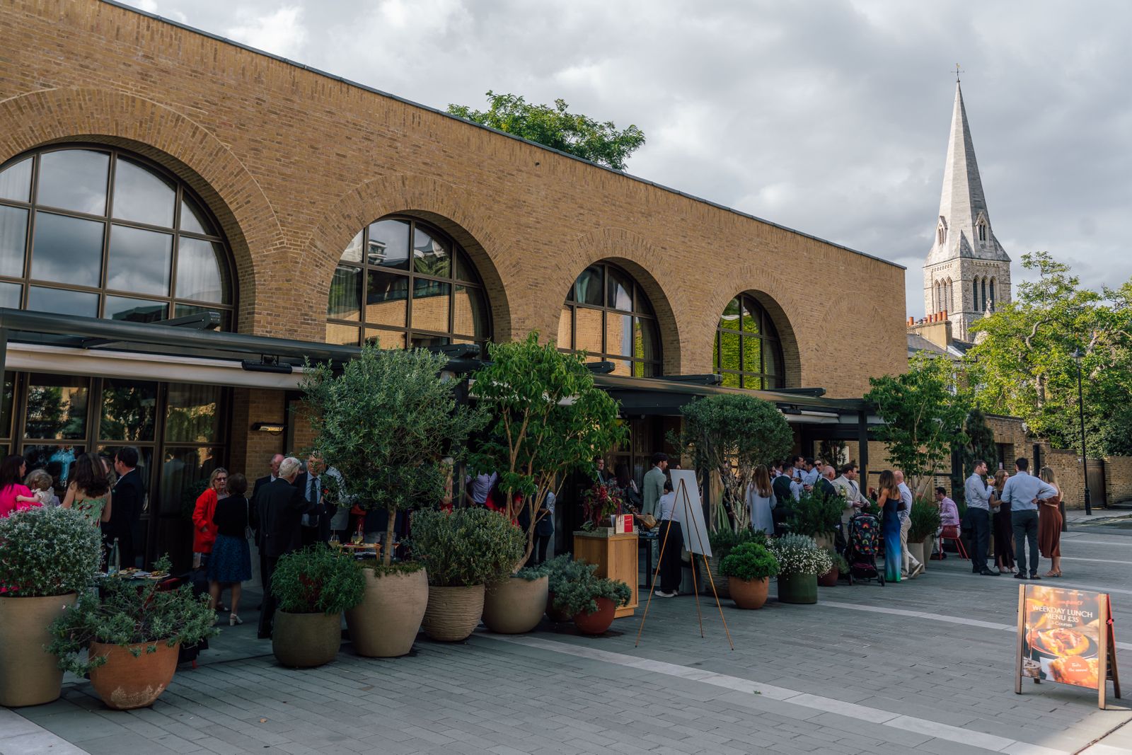 Sunlit indoor dining space with patterned benches, wooden tables, and an olive tree surrounded by lush greenery