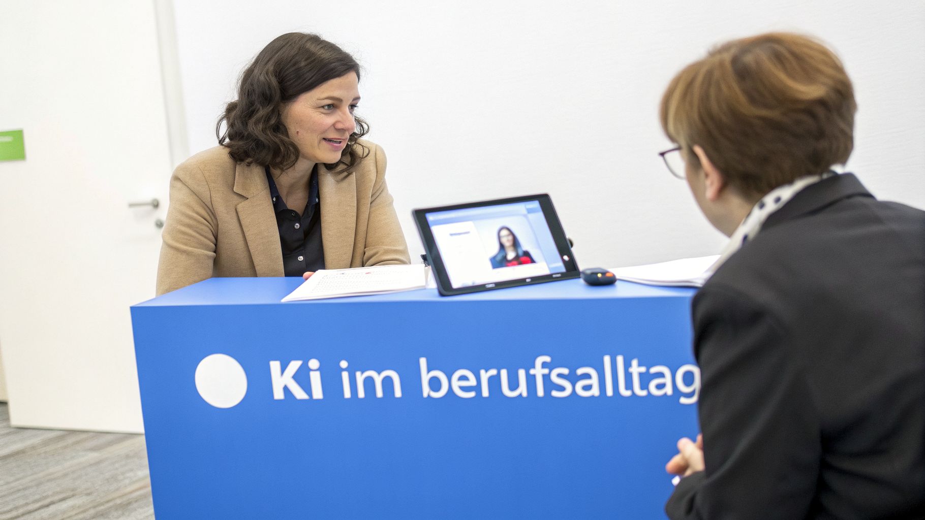 Zwei Frauen unterhalten sich an einem Tisch mit Tablet, das einen Videoanruf zeigt. Thema: Ki im Berufsalltag.
