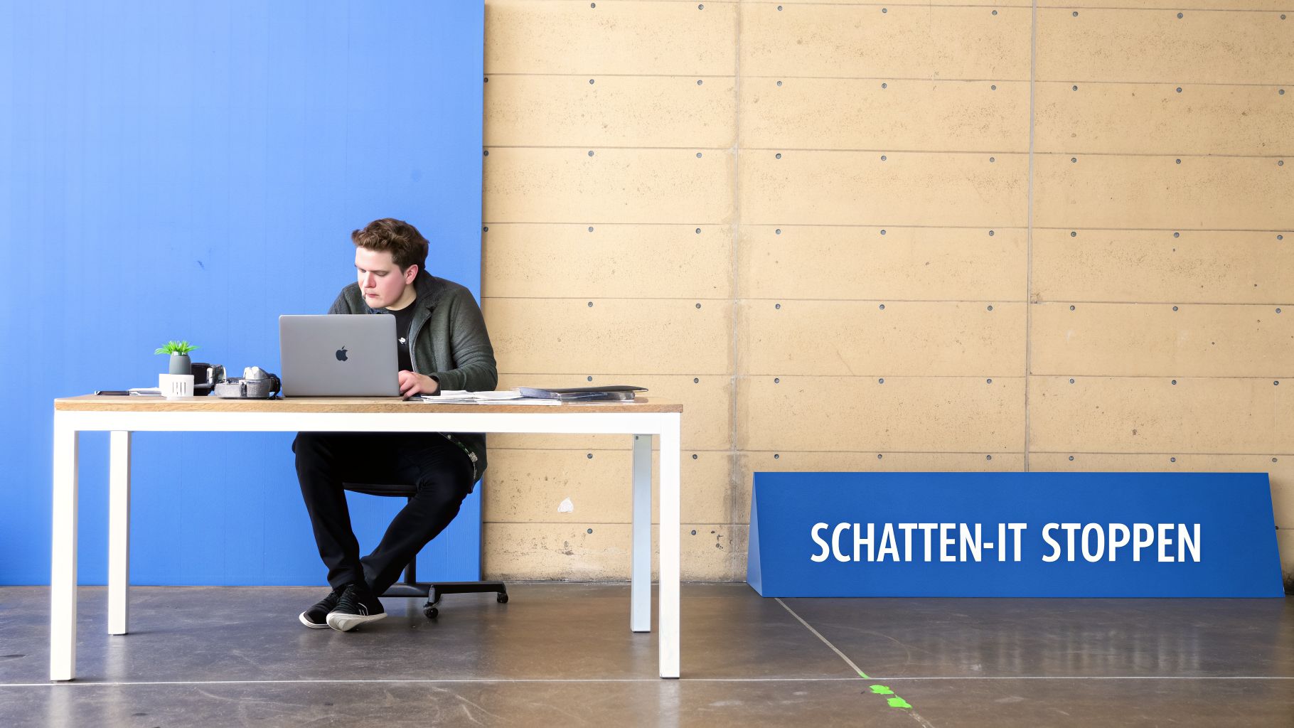 Ein Mann arbeitet konzentriert an einem Laptop an einem Schreibtisch, vor einem modernen Hintergrund mit der Aufschrift 'SCHATTEN-IT STOPPEN'.