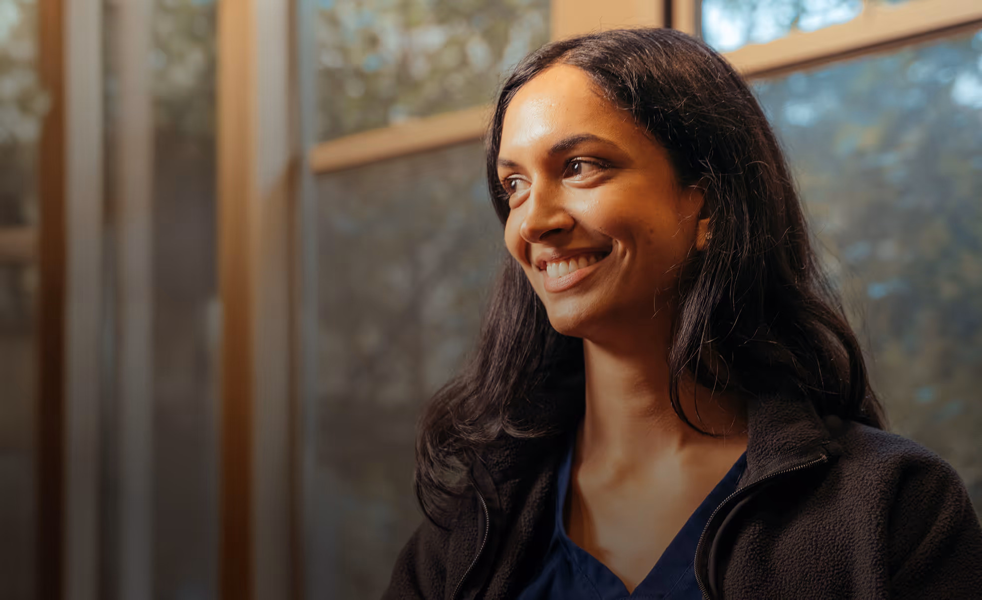 Smiling woman in a dark jacket looking to the side with warm light coming through a window behind her.