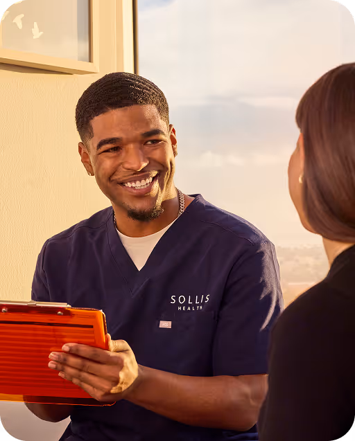 Smiling male healthcare professional in SOLLIS Health scrubs holding an orange clipboard talking to a seated woman.