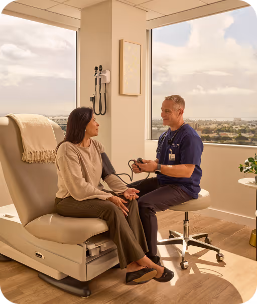 A male healthcare professional in navy scrubs taking blood pressure of a seated female patient in a bright medical office with large windows.