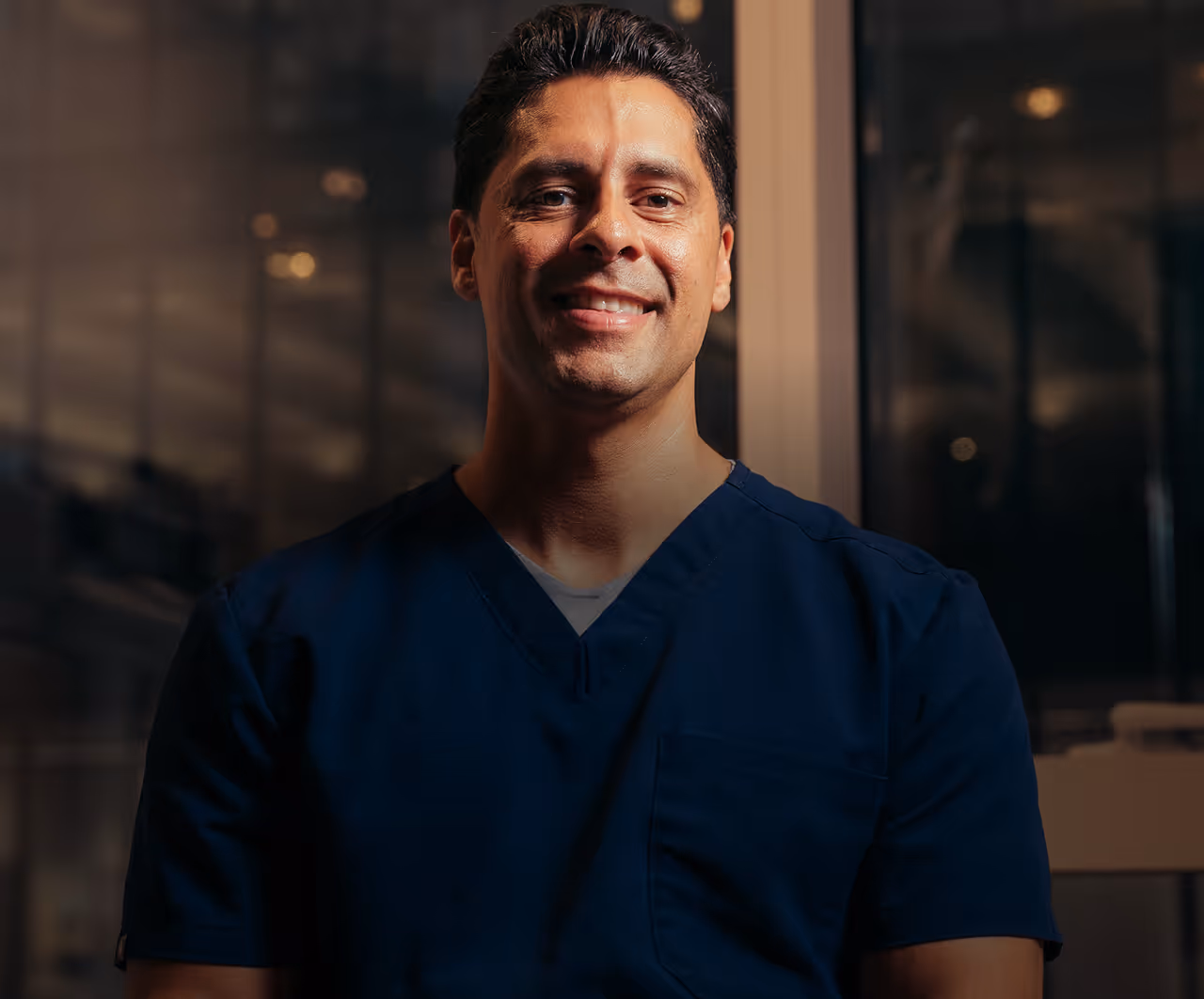 Smiling man with dark hair wearing navy blue medical scrubs in a dimly lit indoor setting.