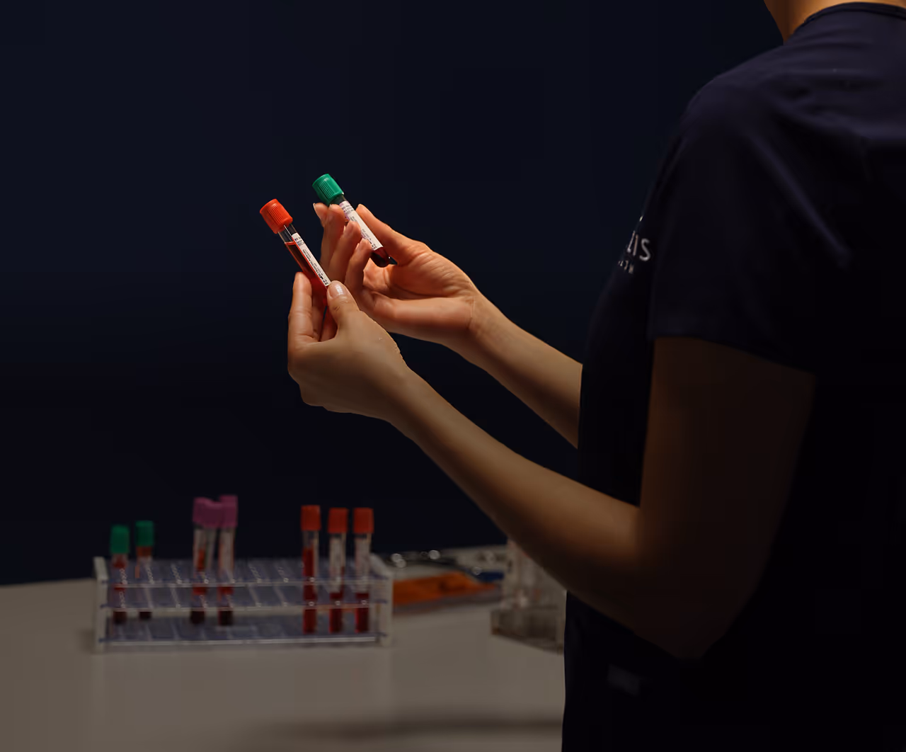 Person holding two blood sample vials with red and green caps, with more vials in a rack on a table.