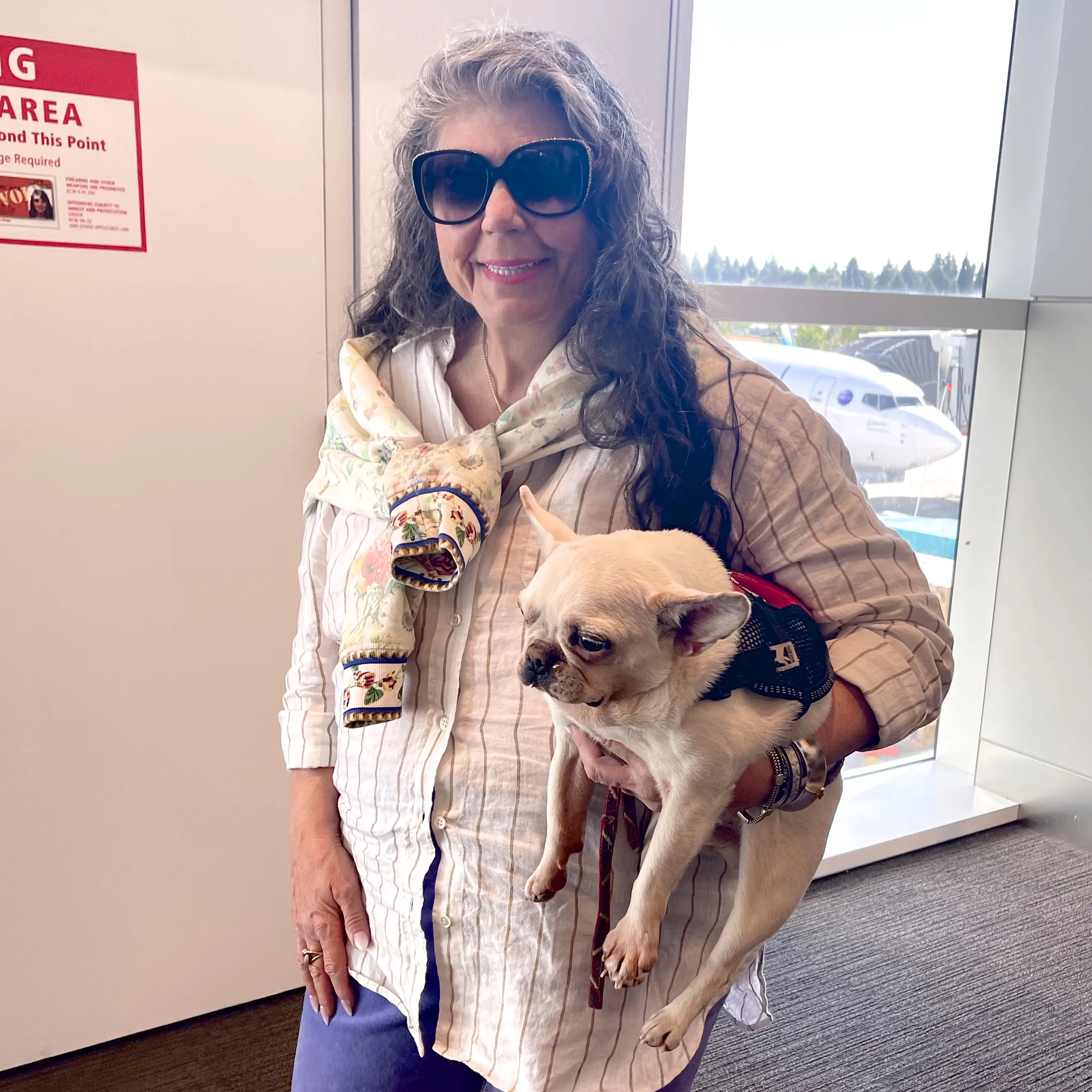 Anahata Graceland standing in an airport while holding a Small Royal Frenchel service dog.