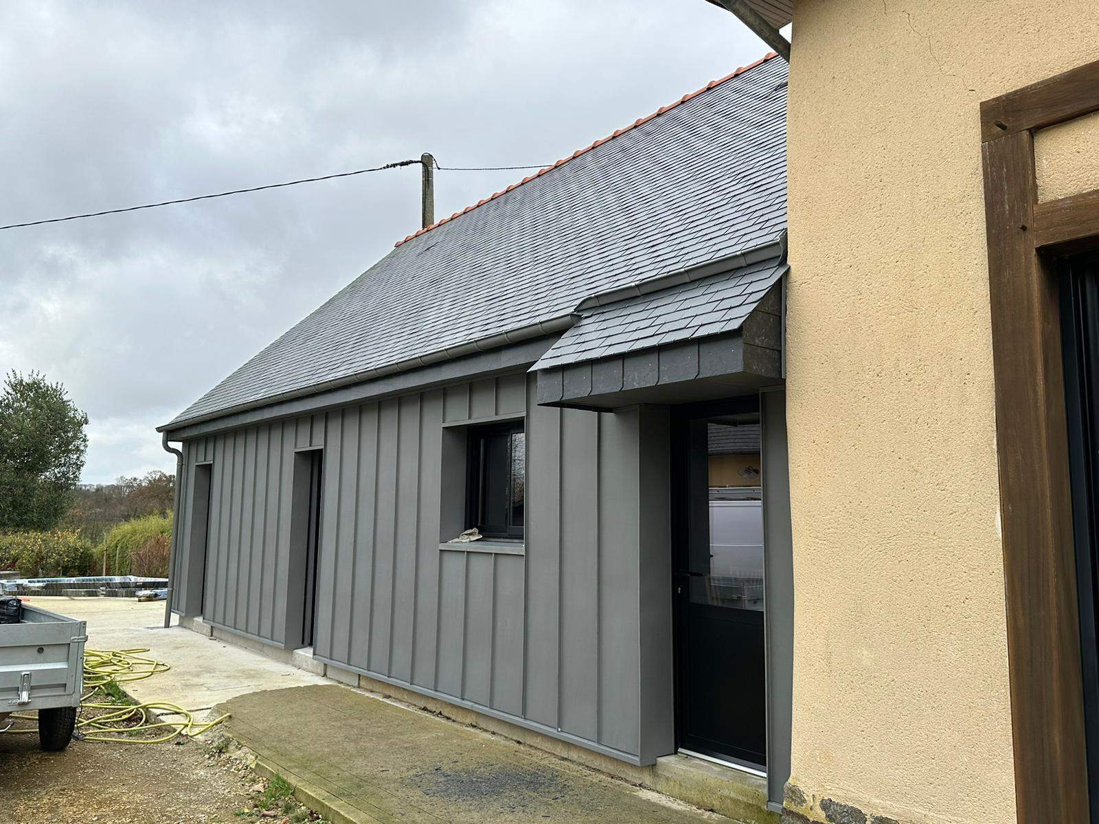 Façade en zinc quartz avec fenêtre, porte et auvent en ardoise contre la maison existante à La Mézière