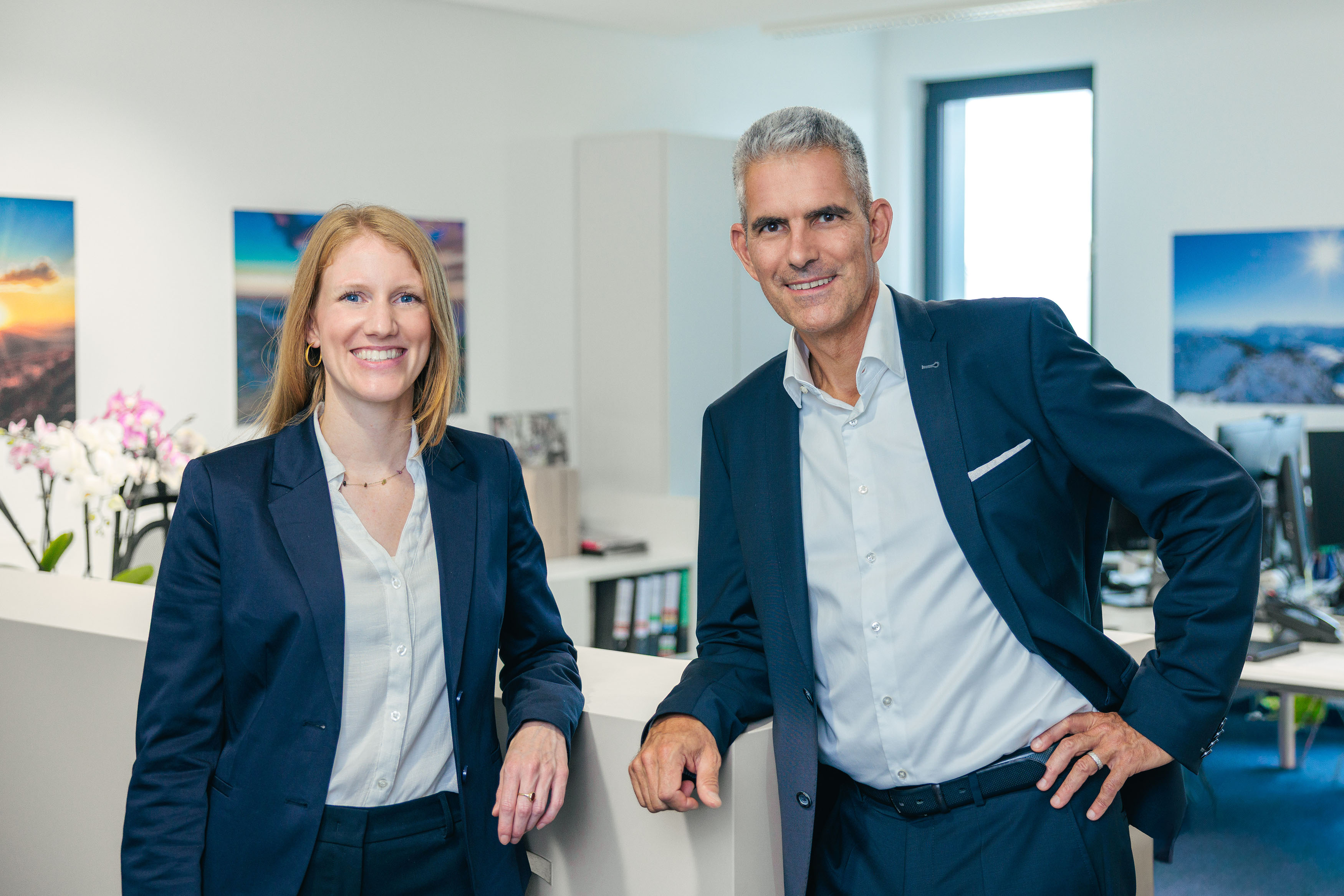 Zwei Personen in Businesskleidung stehen nebeneinander und lächeln.