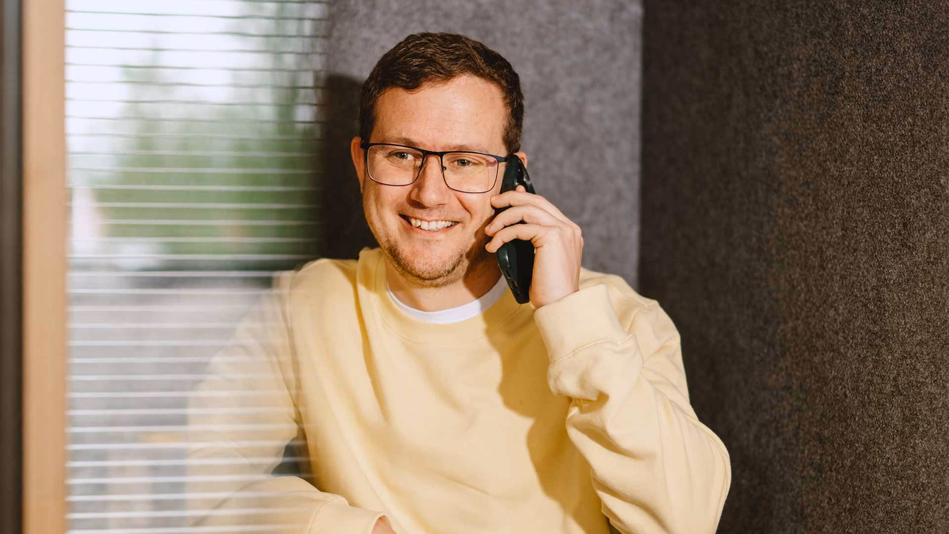 Man met bril en gele trui lacht terwijl hij op een telefoon spreekt in een hoek met raam.