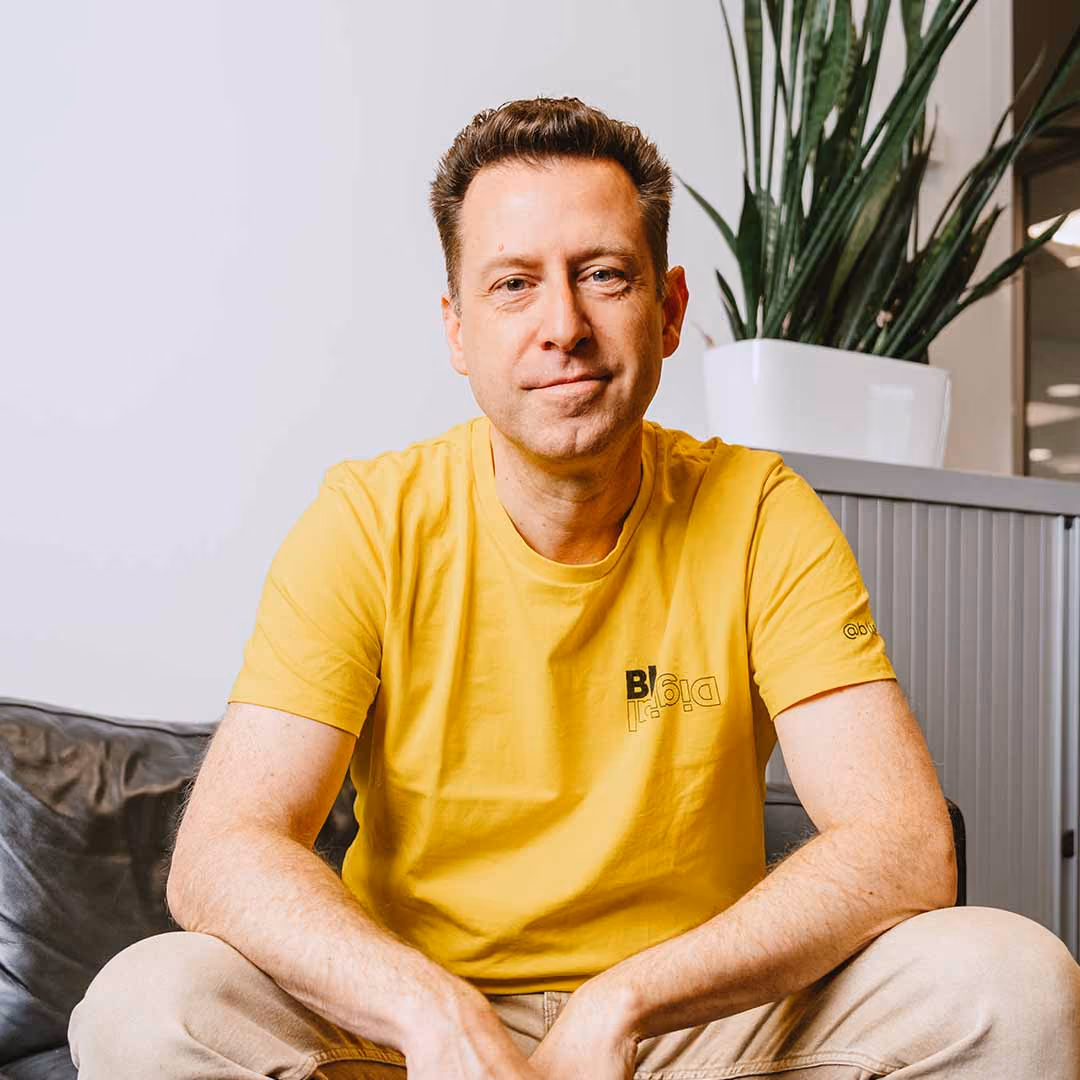 Man in geel T-shirt met Blis Digital logo zit ontspannen op een bank met een plant op de achtergrond.