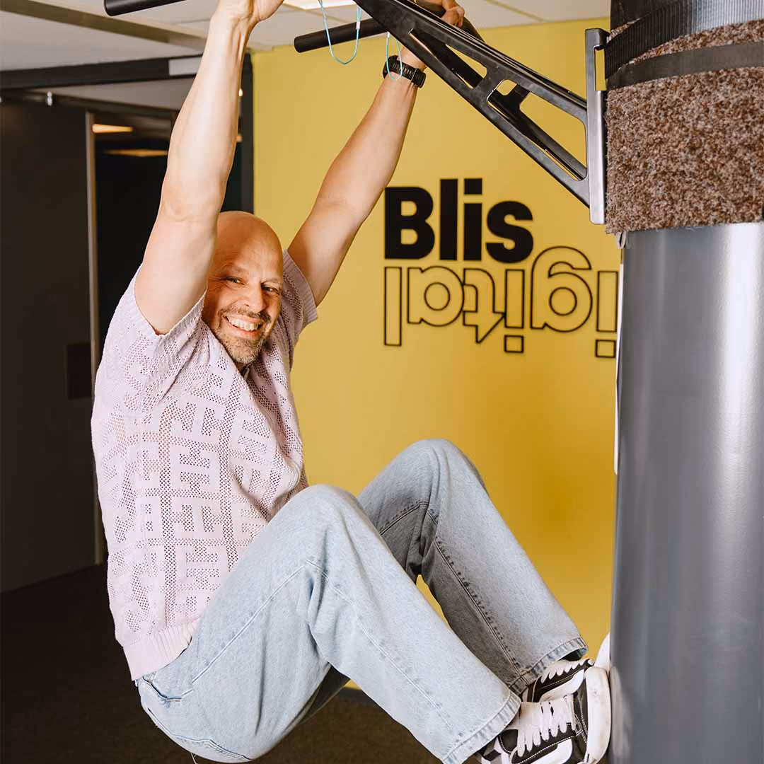 Man lacht en hangt aan een metalen stang aan een gele muur met tekst 'Blis Digital' in een binnenruimte.