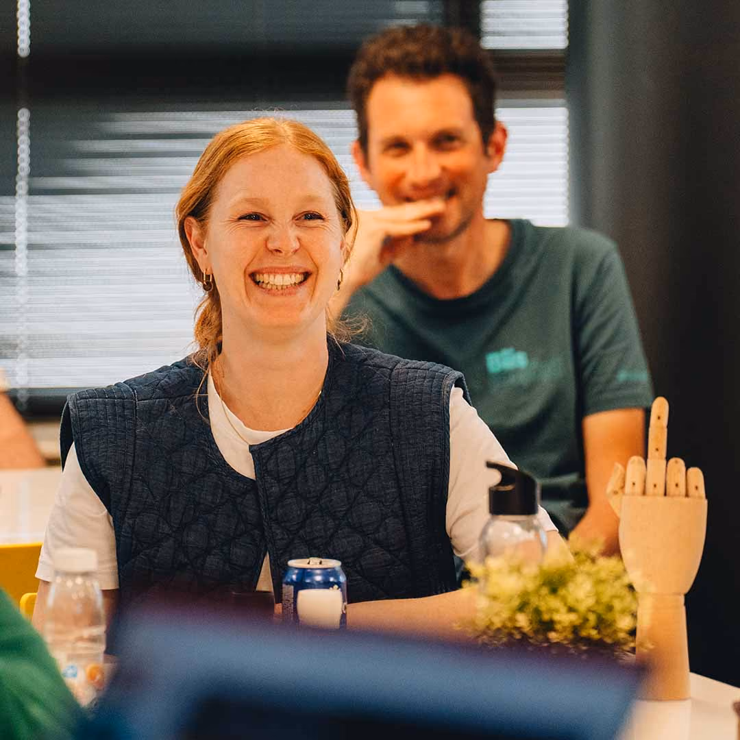 Vrouw lacht zittend aan tafel met drankje, man lacht op achtergrond.