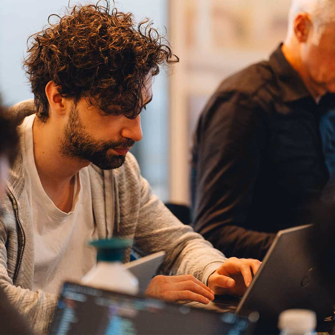 Man met krullend haar werkt geconcentreerd op een laptop in een binnenruimte.