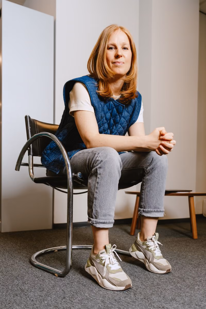 Vrouw met rood haar zit ontspannen op een zwarte stoel in een lichte kamer met casual kleding.
